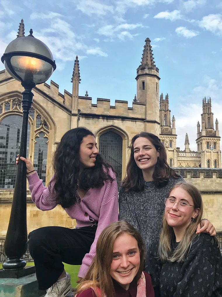 Four people smiling, posing near a historical building with spires, under a bright sky and next to a streetlamp.