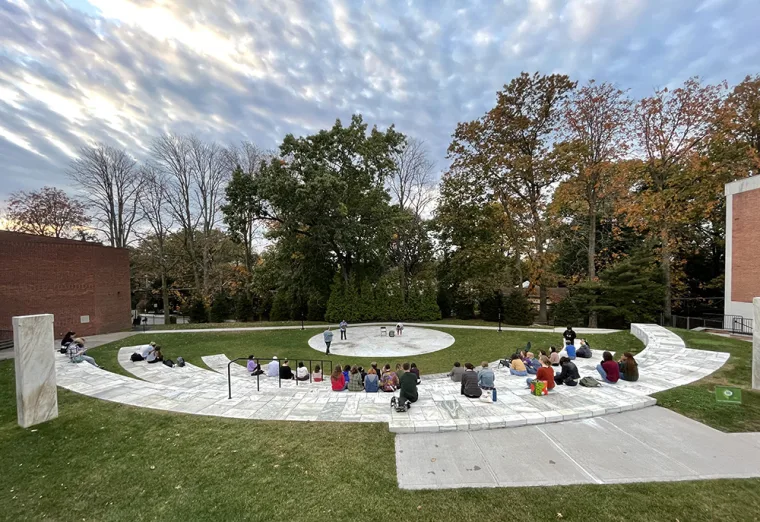 Outdoor amphitheater with a circular seating area where people are gathered, surrounded by trees and buildings under a cloudy sky.