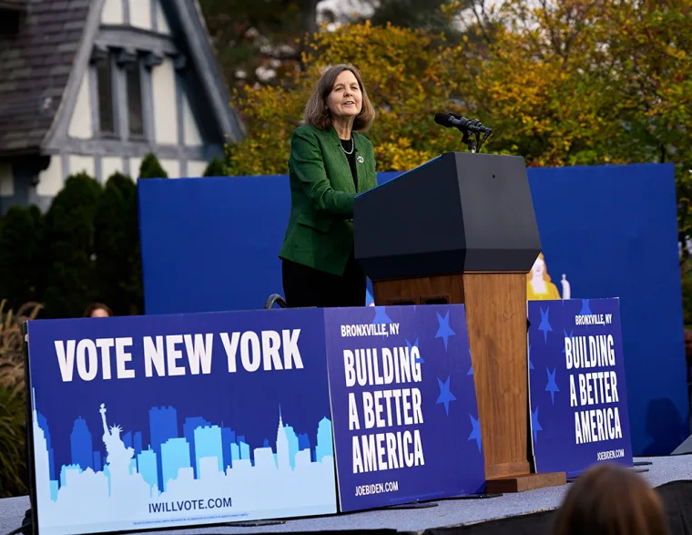 Person speaking at a podium in front of campaign signs that read, "Vote New York" and "Building a Better America," with trees and a house in the background.