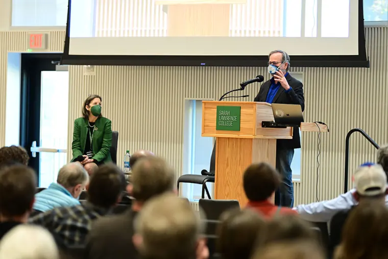Speaker at a podium with Sarah Lawrence College sign, wearing a mask. Audience listens. Another person sits nearby, also masked.