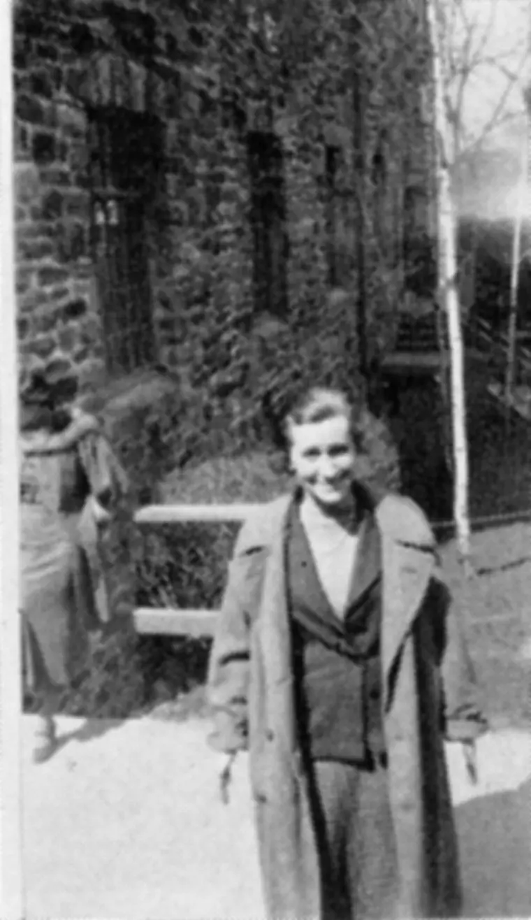 Person in a coat standing outside next to a stone building and wooden fence, with another person walking in the background.