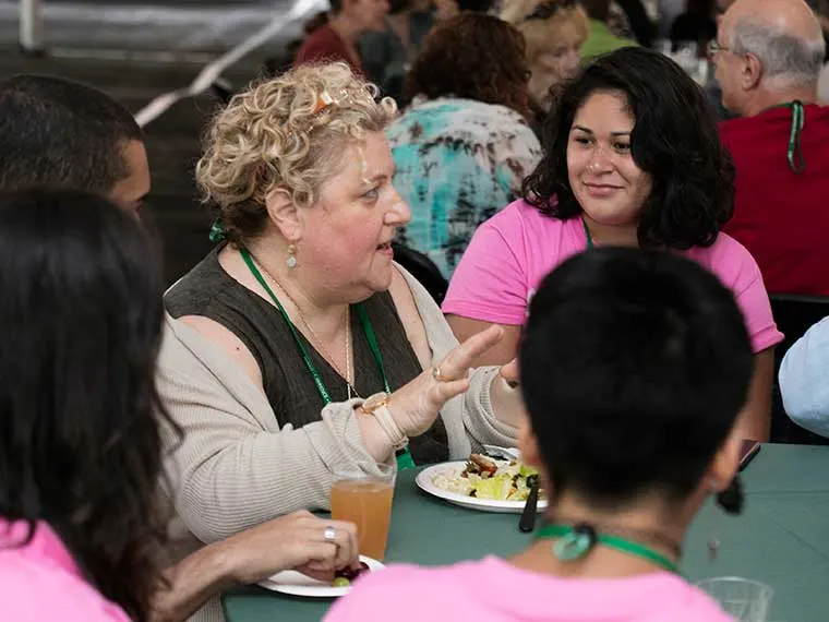 Person gesturing while talking with others at a table, with plates of food and drinks visible.