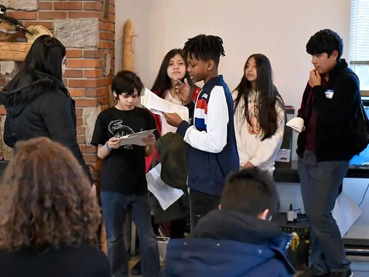 Group of individuals gathered indoors, with some reading papers and others observing, near a brick wall and a large window.