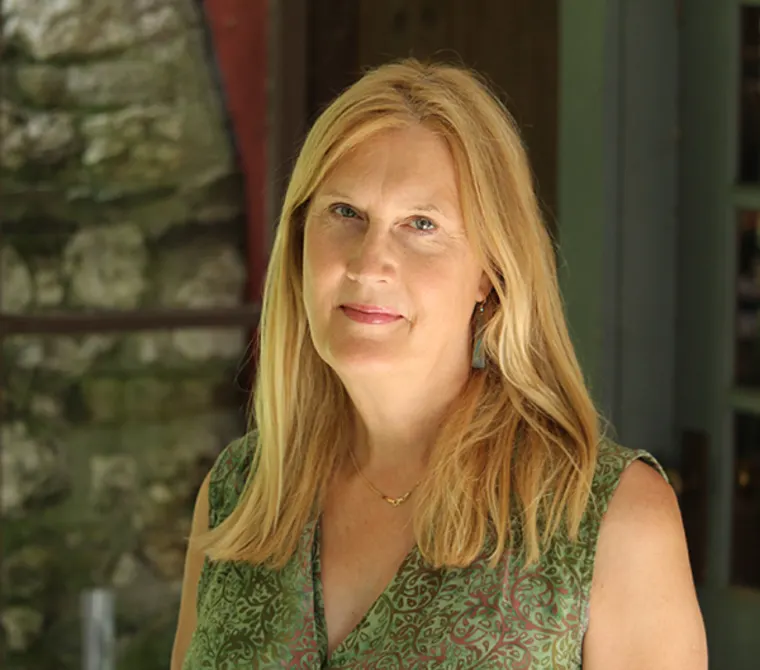 Person with long blond hair in a green patterned top, standing outdoors with a blurred stone wall in the background.