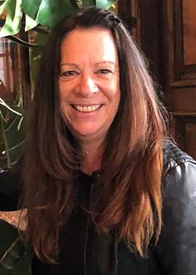Smiling person with long brown hair wearing a dark jacket stands indoors, partially obscured by leaves in the background.