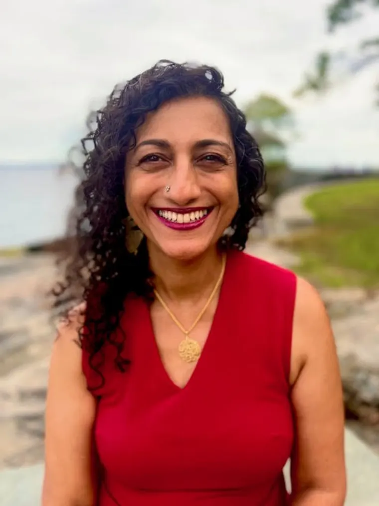 Person with curly hair and red top smiles outdoors by a path and water in the background.