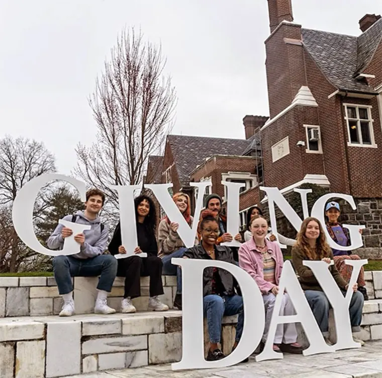 Group of people sitting on steps holding large letters spelling "GIVING DAY," with a brick building and trees in the background.