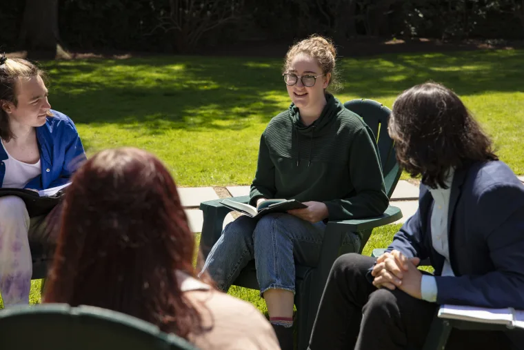 People sitting in a circle on a lawn, talking and holding books, with trees in the background.