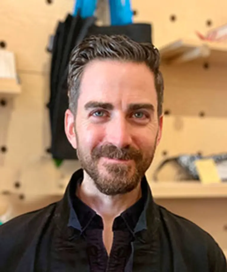 Person with short hair and beard smiling, wearing a dark jacket in front of a wall with shelves and various items.