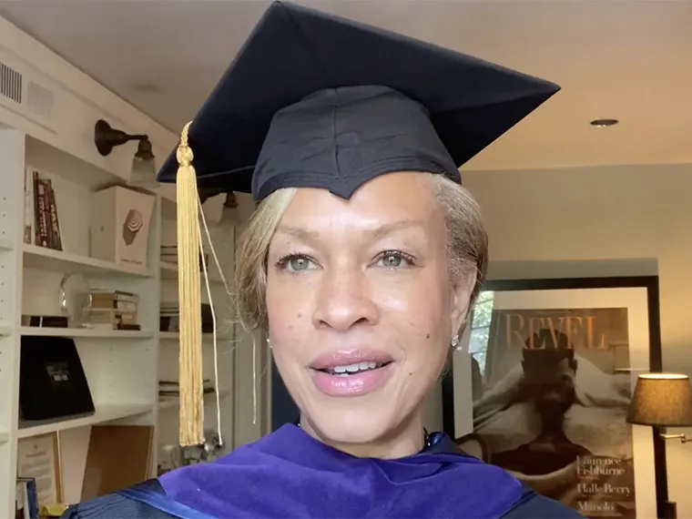 Person wearing a graduation cap and gown, smiling in a room with shelves and a poster in the background.