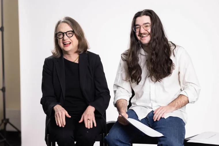 Two people sitting and smiling, holding papers. One is wearing a black outfit and glasses, the other a white shirt and jeans with long hair.