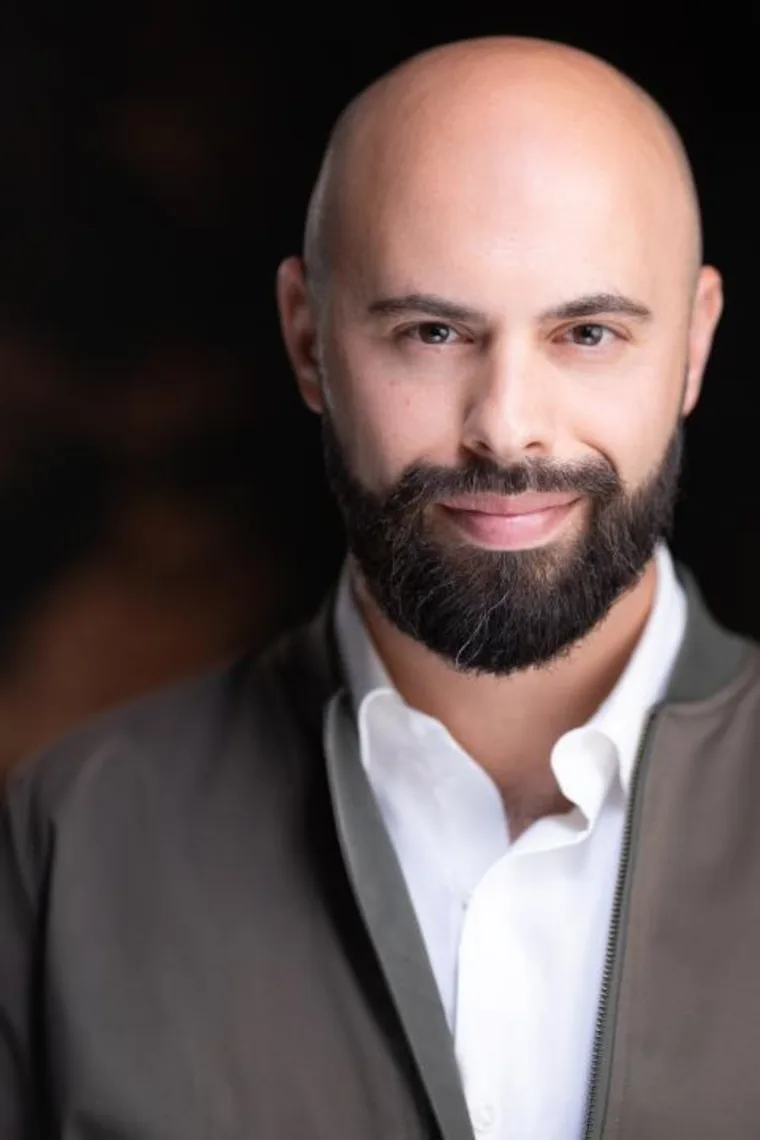 A person with a bald head and beard, wearing a white shirt and dark jacket, smiles softly against a dark background.