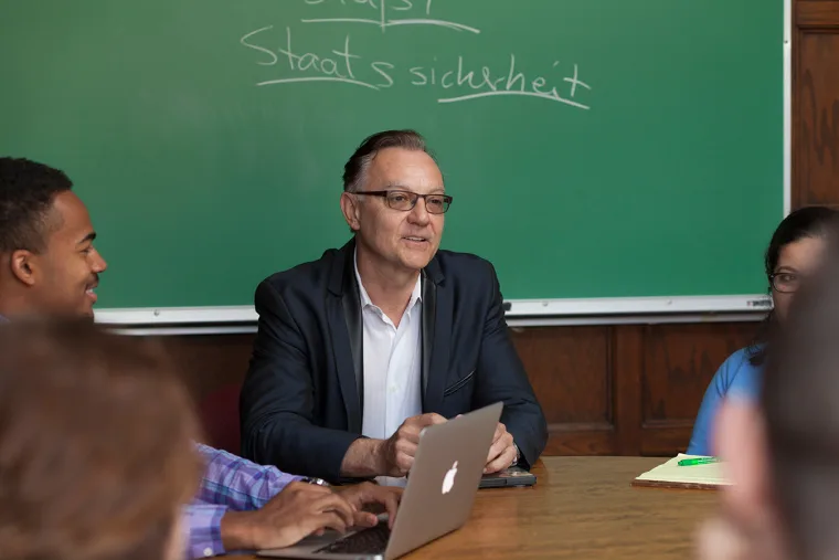 Person speaking at a table with a laptop, surrounded by others, in front of a green chalkboard with writing.