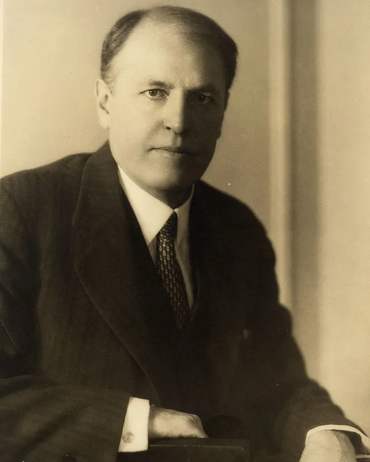 Person in a suit with short hair, posed in a formal portrait, looking directly at the camera with a neutral expression. Sepia tone.