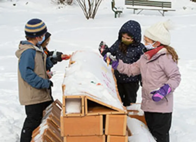 Four children wearing winter clothing and masks paint a snowy structure outdoors. A bench and bare trees are in the background.
