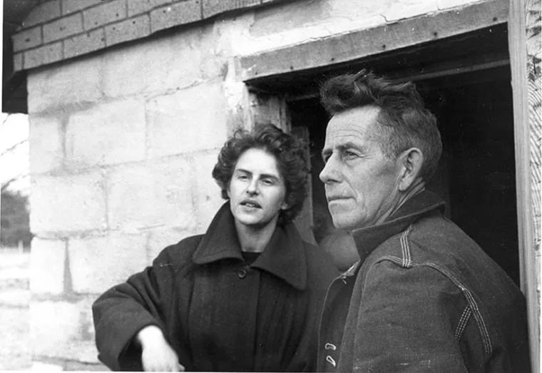 Two people standing outside a brick building, one looking directly at the camera and the other gazing into the distance.
