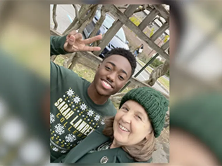 Two people outdoors taking a selfie. One is smiling and giving a peace sign, wearing a "Sarah Lawrence" sweater. The other is wearing a green hat.