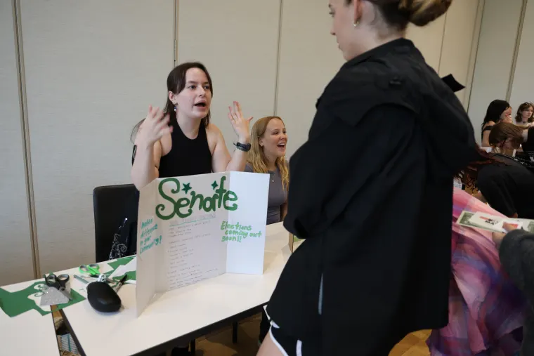 Person at a table with a "Senate" sign, talking to another person. Papers and mouse on the table. Others in the background.