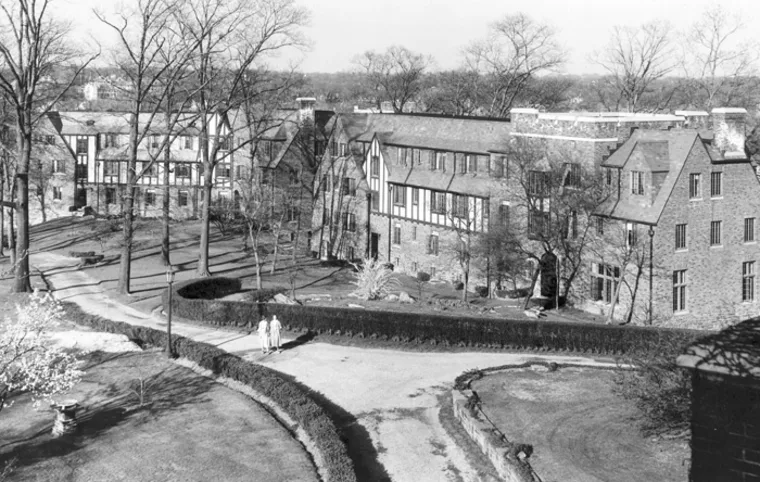 Historic black and white image of a large stone building with a pitched roof, surrounded by bare trees and a winding path.