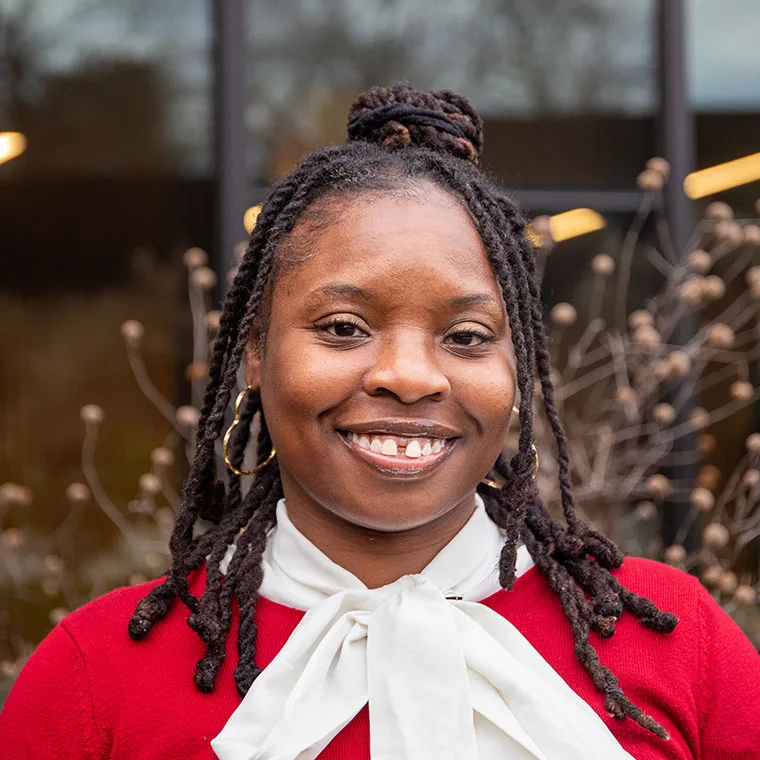 Person smiling, wearing a red sweater and white bow tie, with braided hair, standing outdoors with blurred plants and a building in the background.