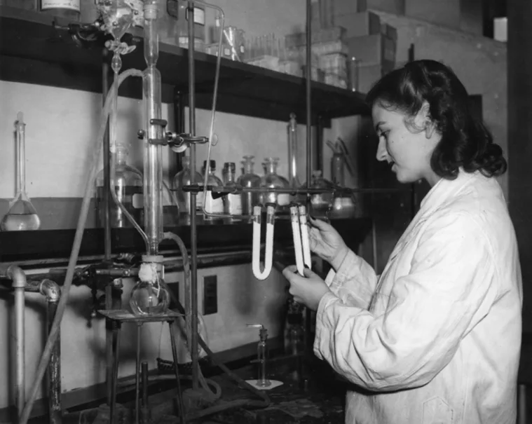 Person in a lab coat working with test tubes in a laboratory setup. Shelves with glassware in the background.