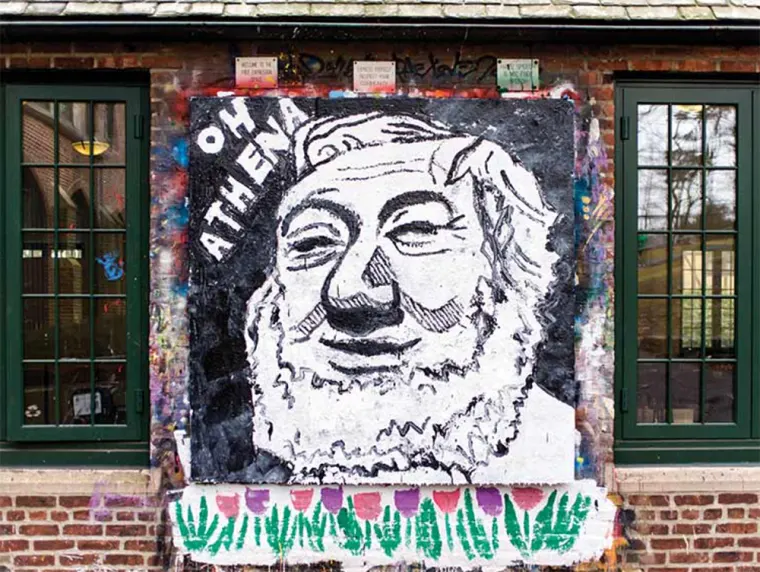 Street art on a brick wall depicts a bearded face with "Oh Athena" written beside it. Two windows with green frames flank the artwork.