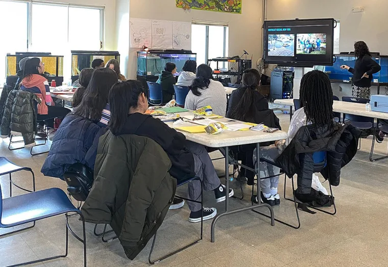 Classroom with students seated at tables watching a presenter at a large screen. Desks have books and snacks. Aquariums line the walls.