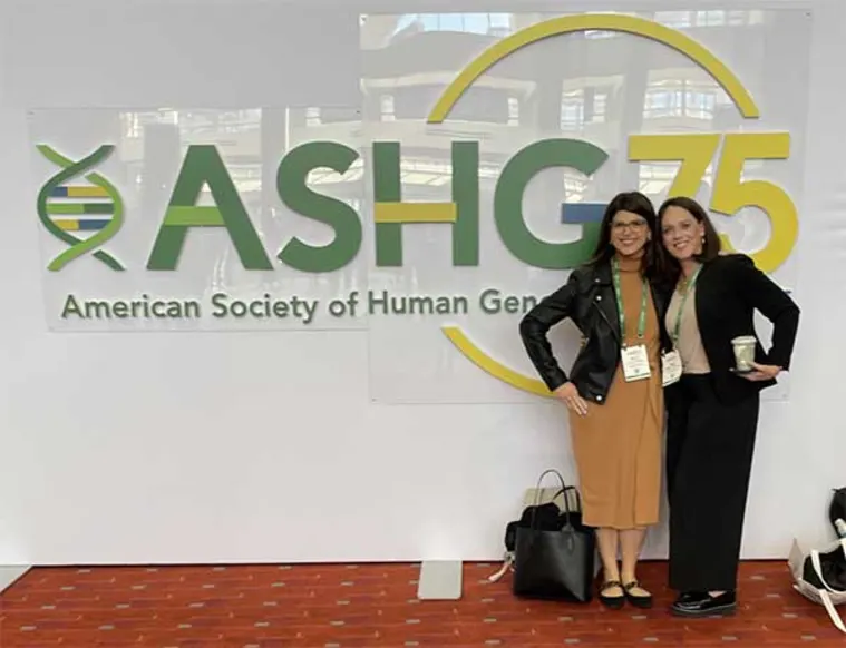 Two people stand smiling in front of an ASHG 75 sign at an event, wearing name badges and conference attire.