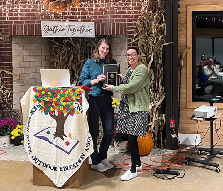 Two people stand by a podium draped with a decorated cloth; one holds a plaque in a room with a "Gather Together" sign.
