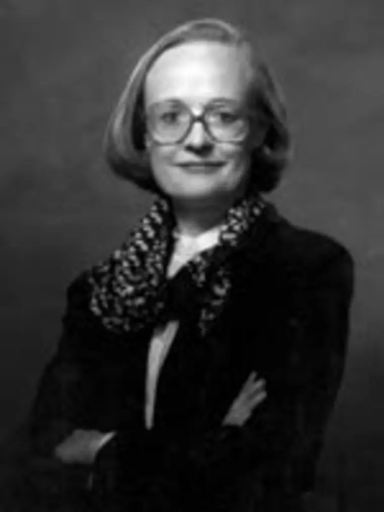 Person with short hair and glasses wearing a dark suit and patterned scarf, arms crossed, against a plain background.