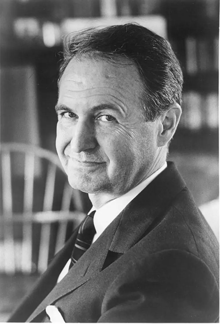 Smiling person in a suit, looking towards the camera with a blurred background.