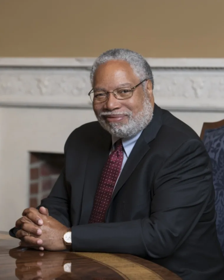 Person with gray hair and beard in a suit and red tie, seated at a wooden table, hands clasped, in a room with beige walls and white molding.