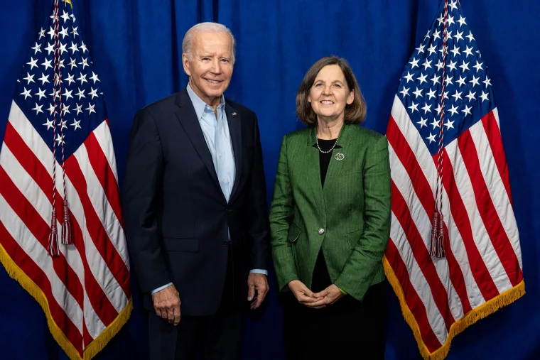 Two people standing and smiling in front of a blue backdrop with two American flags.