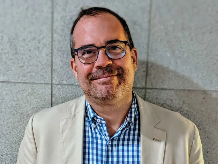 Person smiling, wearing glasses and a checkered shirt with a beige jacket, standing against a tiled wall.