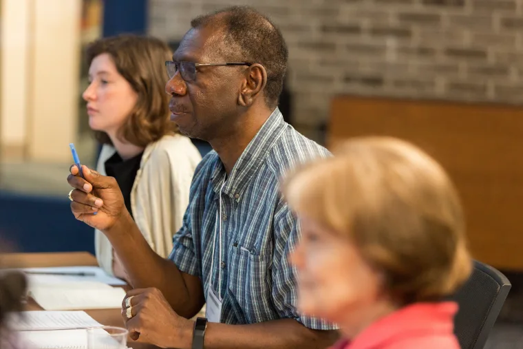 Three people seated at a table, listening attentively. The person in focus holds a pen, appearing engaged in a discussion or meeting.