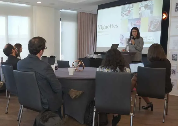 Person presenting in front of a projector screen labeled "Vignettes" to an audience seated at tables with laptops.
