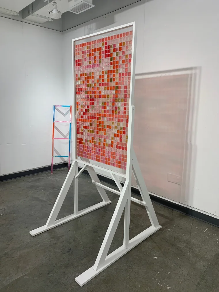 A freestanding artwork with a grid of red and pink squares is displayed on a white frame. A colorful ladder is visible in the background.