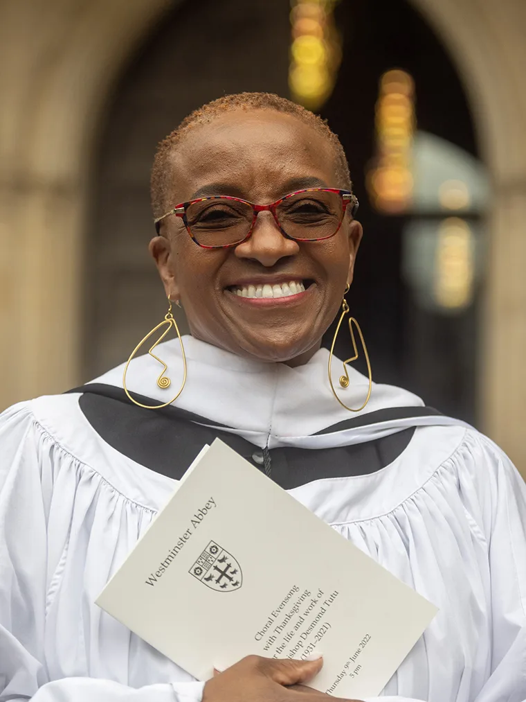 Smiling person in a white robe holds a Westminster Abbey program. They wear red glasses and large, abstract earrings.