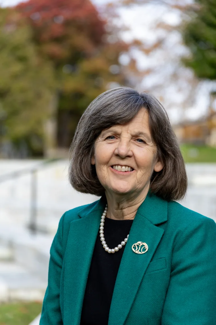 Person with short hair smiling, wearing a green blazer and pearl necklace, standing outdoors with blurred trees in the background.