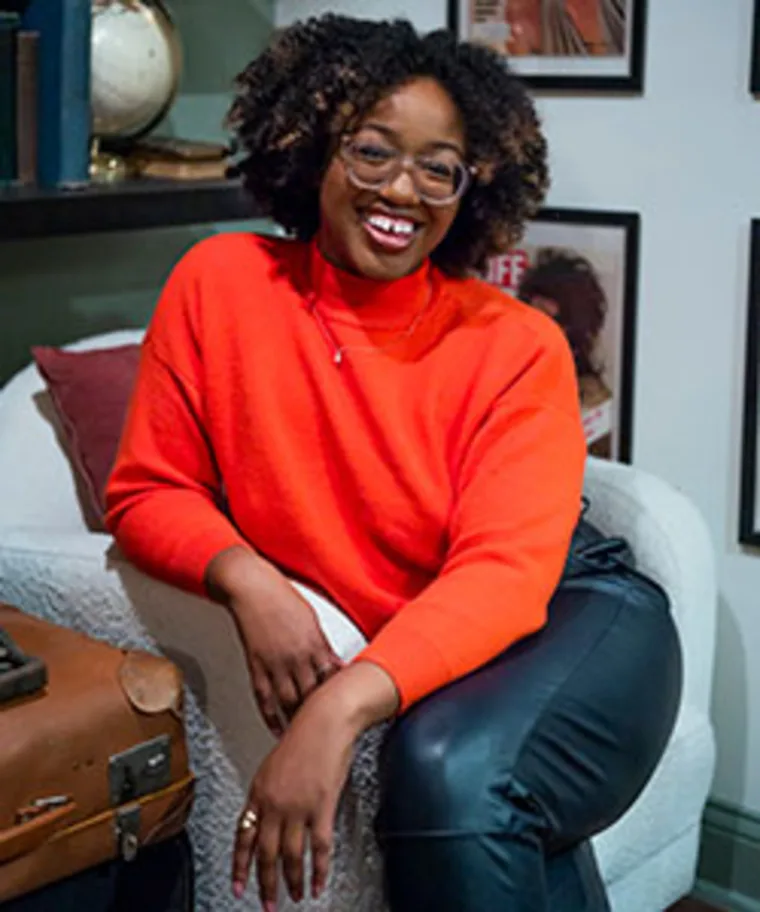 Person with curly hair and glasses, wearing a bright orange sweater and black pants, sitting on a white chair, smiling, with framed photos behind.