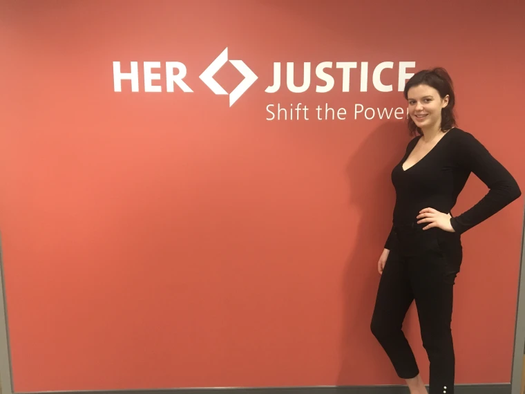 Person standing beside a red wall with "HER JUSTICE Shift the Power" written on it, smiling with one hand on the hip.