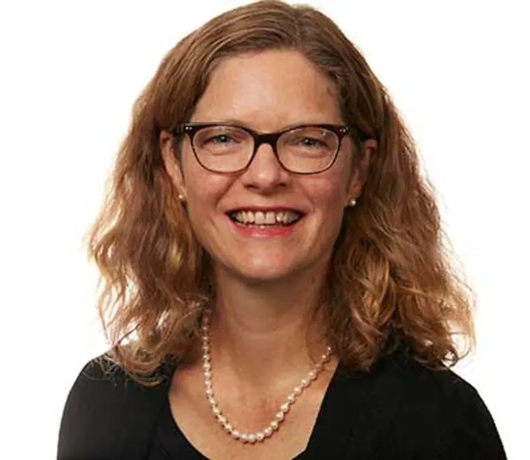 Person with curly hair, glasses, and a pearl necklace smiles against a plain background.