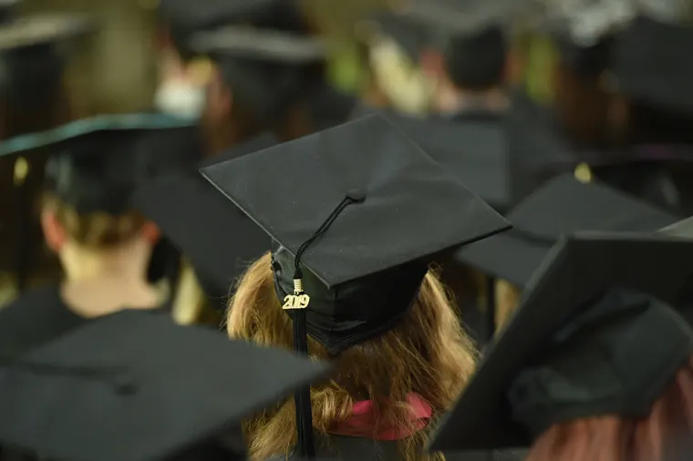 Group of graduates seated at a ceremony, viewed from behind. Focus on a cap with a 2019 tassel.