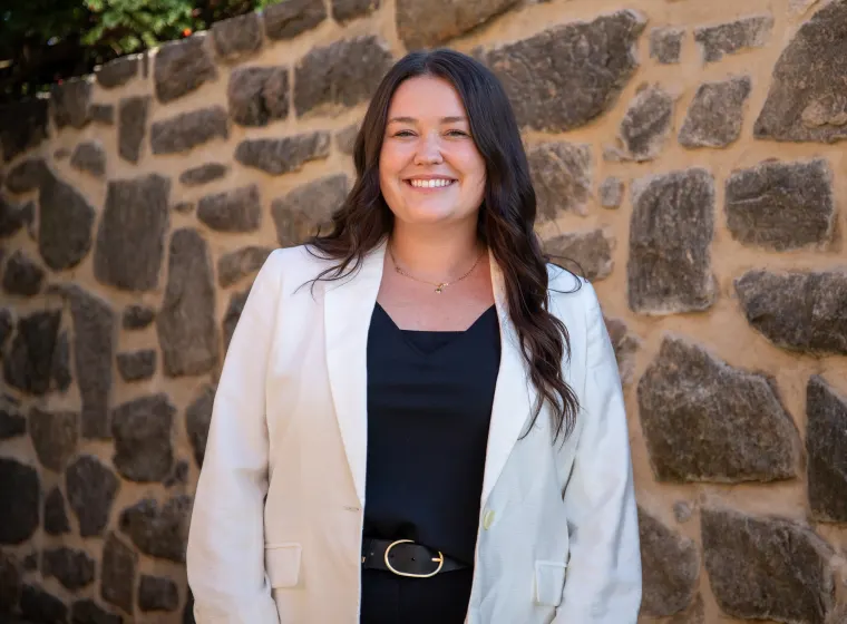 Person with long hair smiling, wearing a white blazer and black top, standing in front of a stone wall.