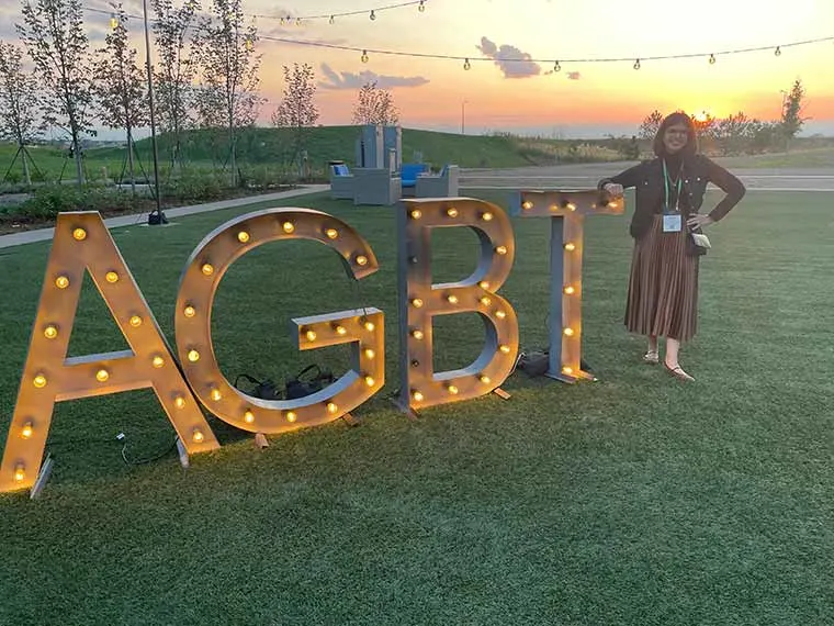 Person standing next to large illuminated letters "AGBT" on grass at sunset, with string lights and distant hills in the background.