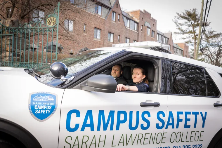 Two individuals in a Campus Safety vehicle at Sarah Lawrence College. A brick building and wrought-iron gate are visible in the background.