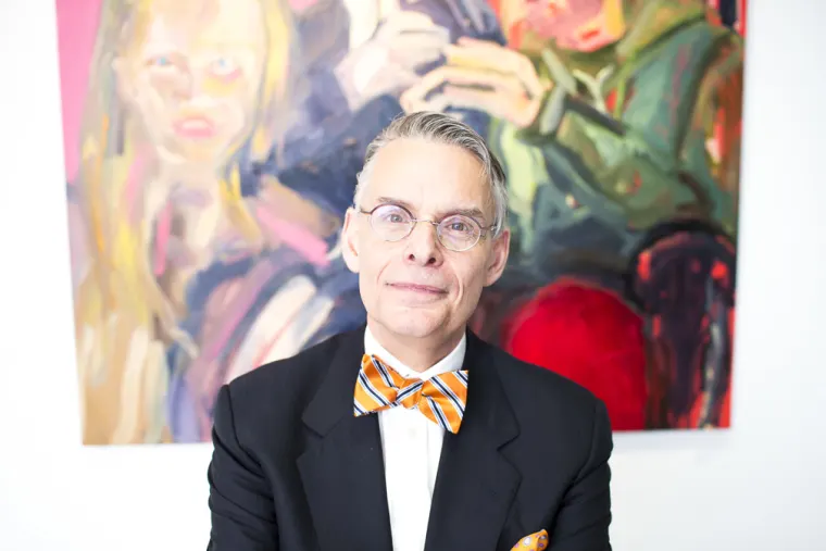 Person wearing glasses and a striped bow tie stands in front of a colorful abstract painting.