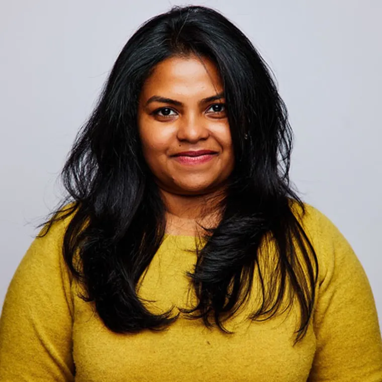 Person with long dark hair wearing a yellow sweater, smiling in front of a plain background.