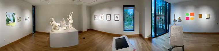 Art gallery with sculptures on a central pedestal, framed works on surrounding walls, wooden floor, and large windows offering natural light.