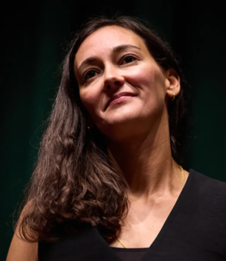 Person with long dark hair and a slight smile, wearing a dark top, looks upward with a thoughtful expression. Dark background.